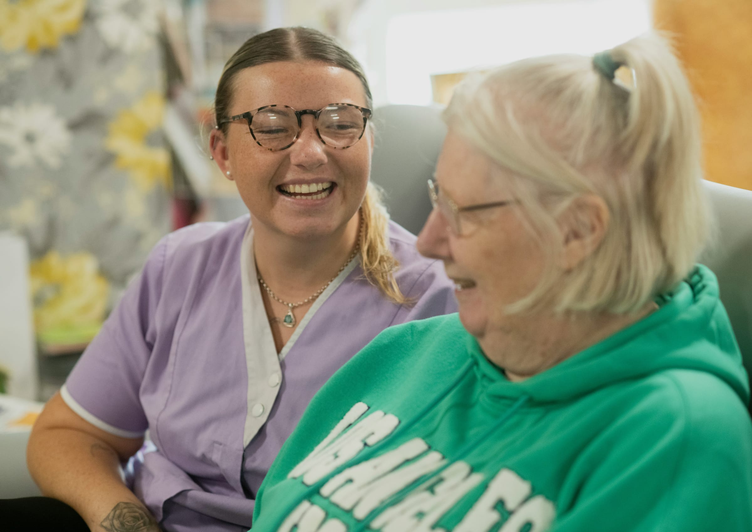 A carer in a lilac tunic laughing with an older woman in her armchair