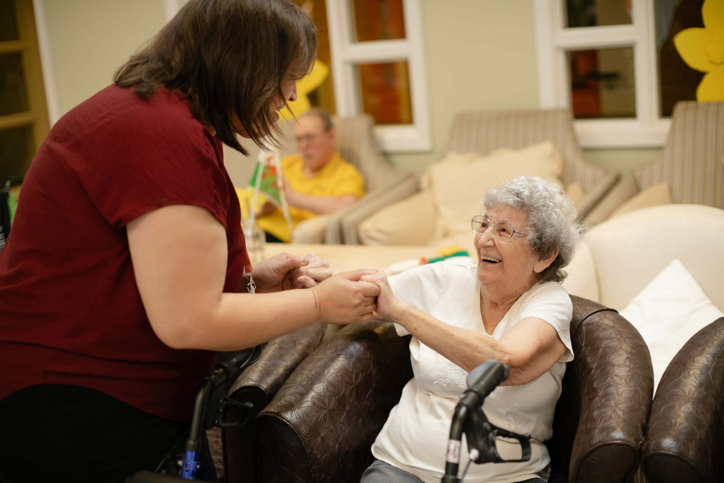 A carer helping a client up from her armchair, holding both her hands